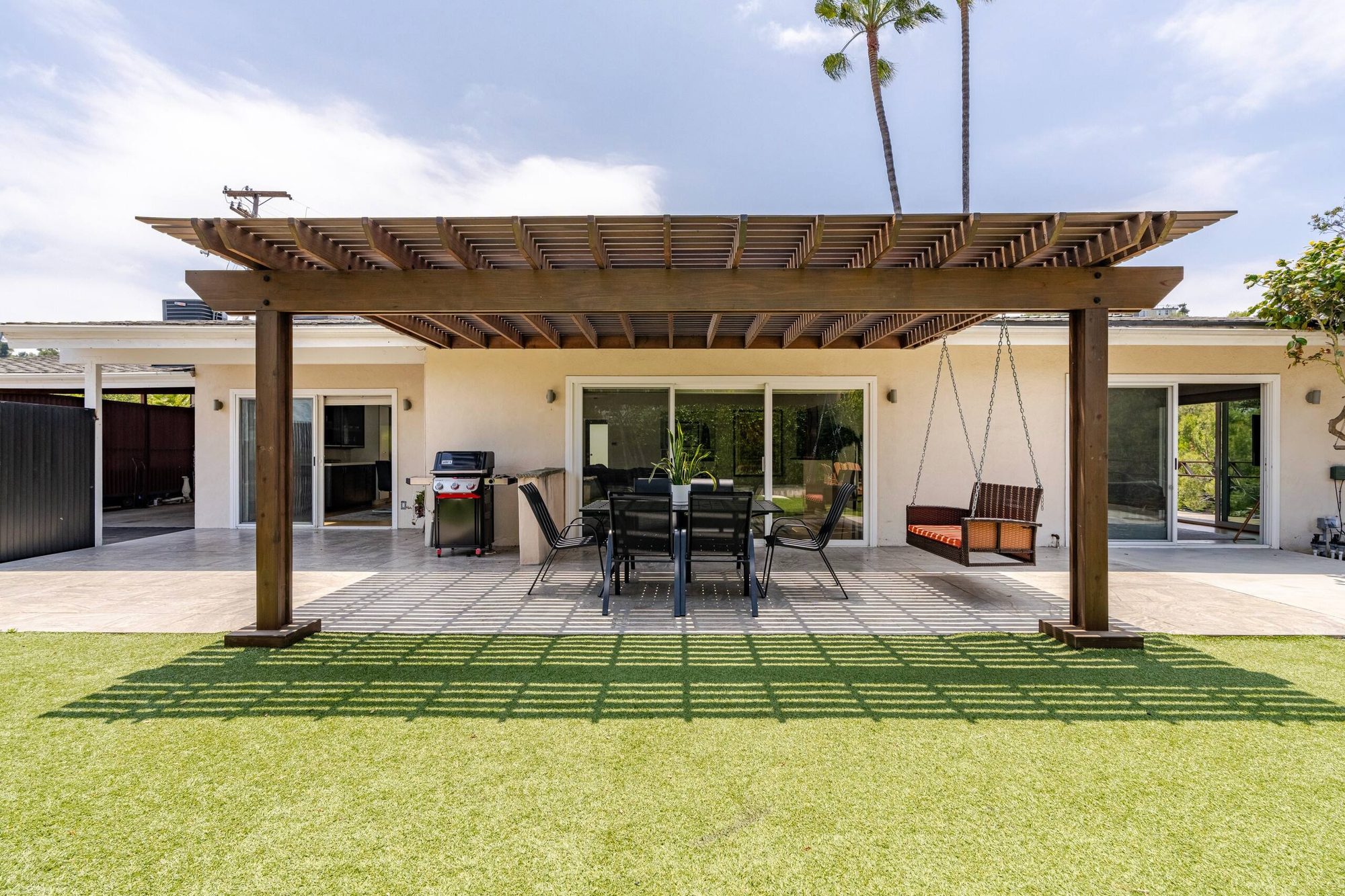 Backyard patio under a wooden pergola with dining table and swinging bench, palm trees overhead
