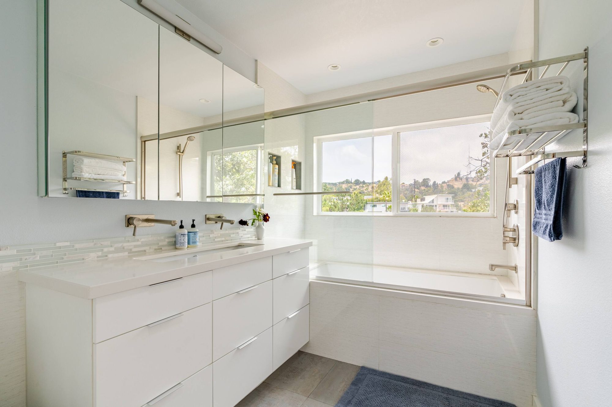 Primary bathroom with long double vanity, mirrored medicine cabinets, and a tub-shower combo beneath a window with a hillside view