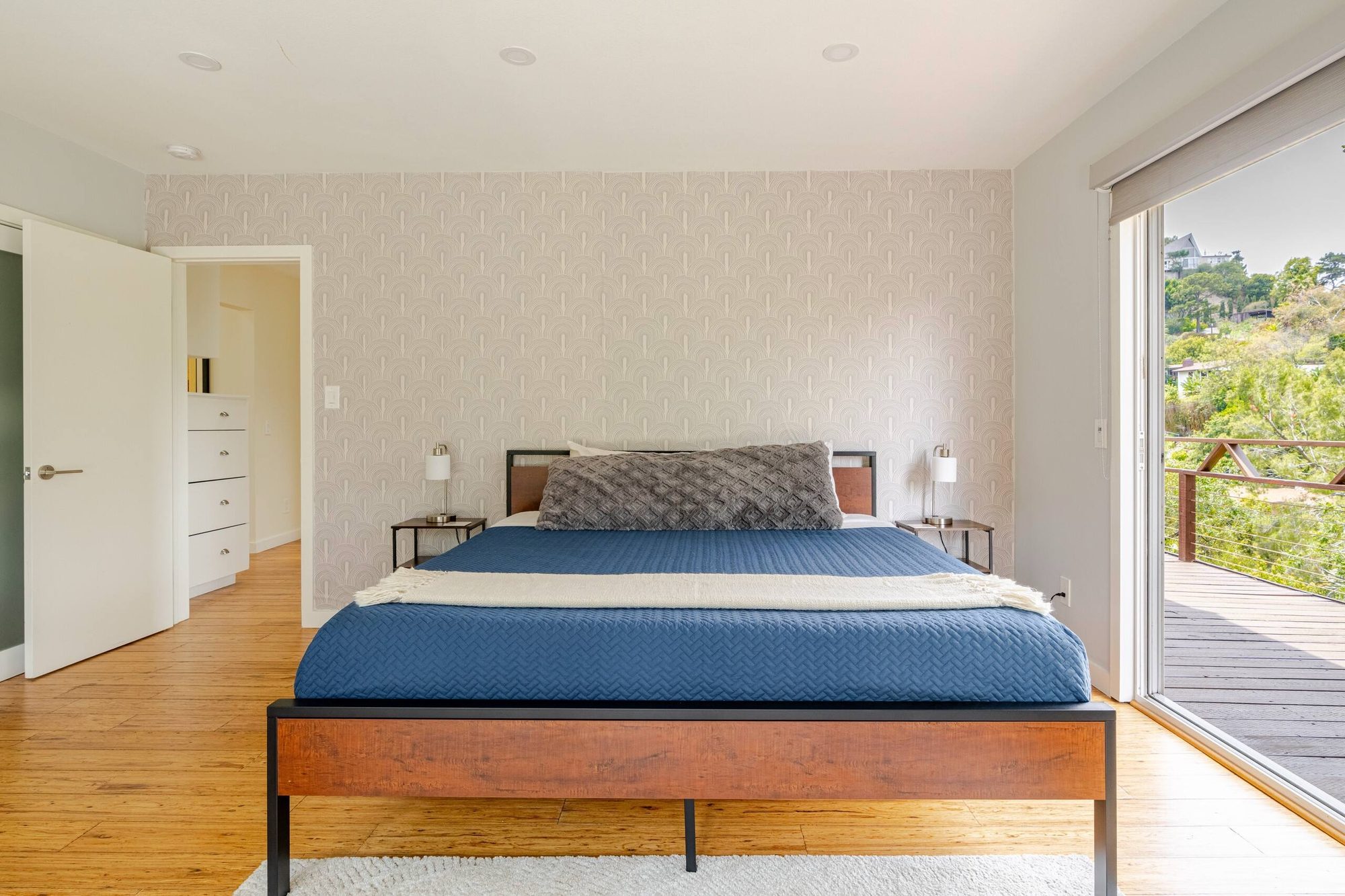 Primary bedroom with patterned wallpaper accent wall, blue bedspread, and sliding door to a private deck